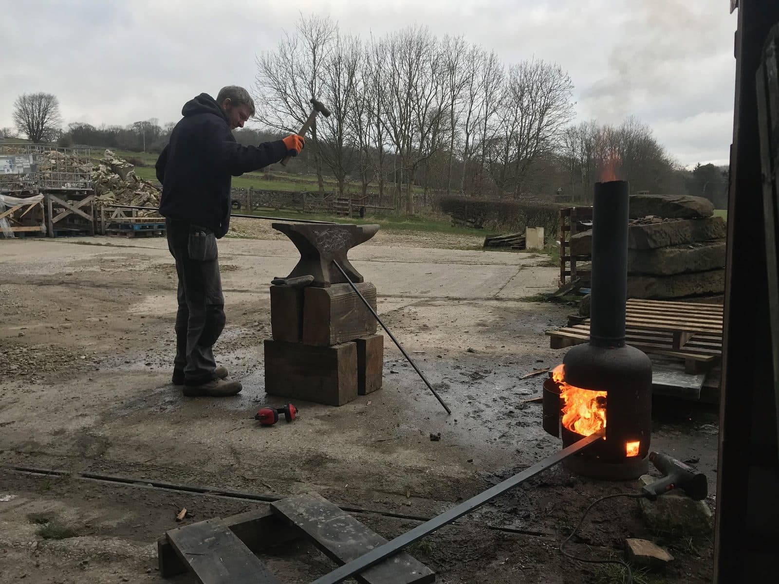Blacksmith hammering on an anvil beside a wood burner in a rural workshop yard