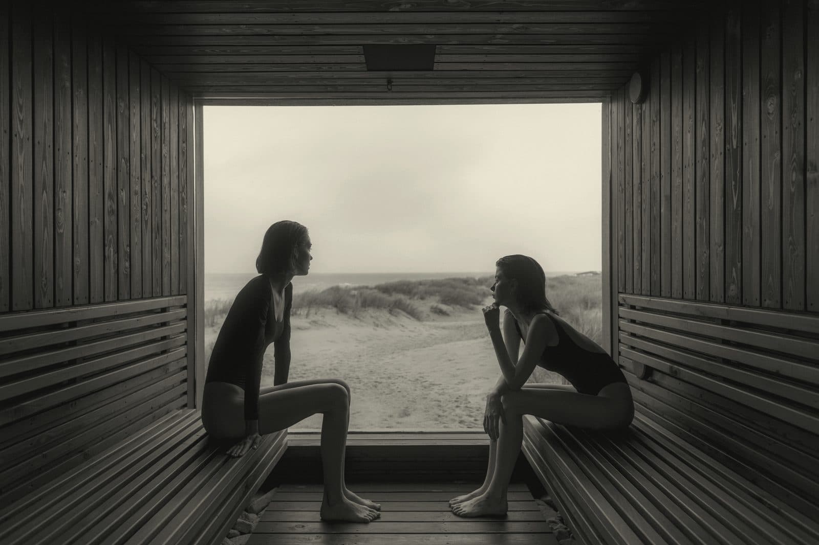Two people sitting face to face inside a timber sauna with open rear wall framing a coastal landscape