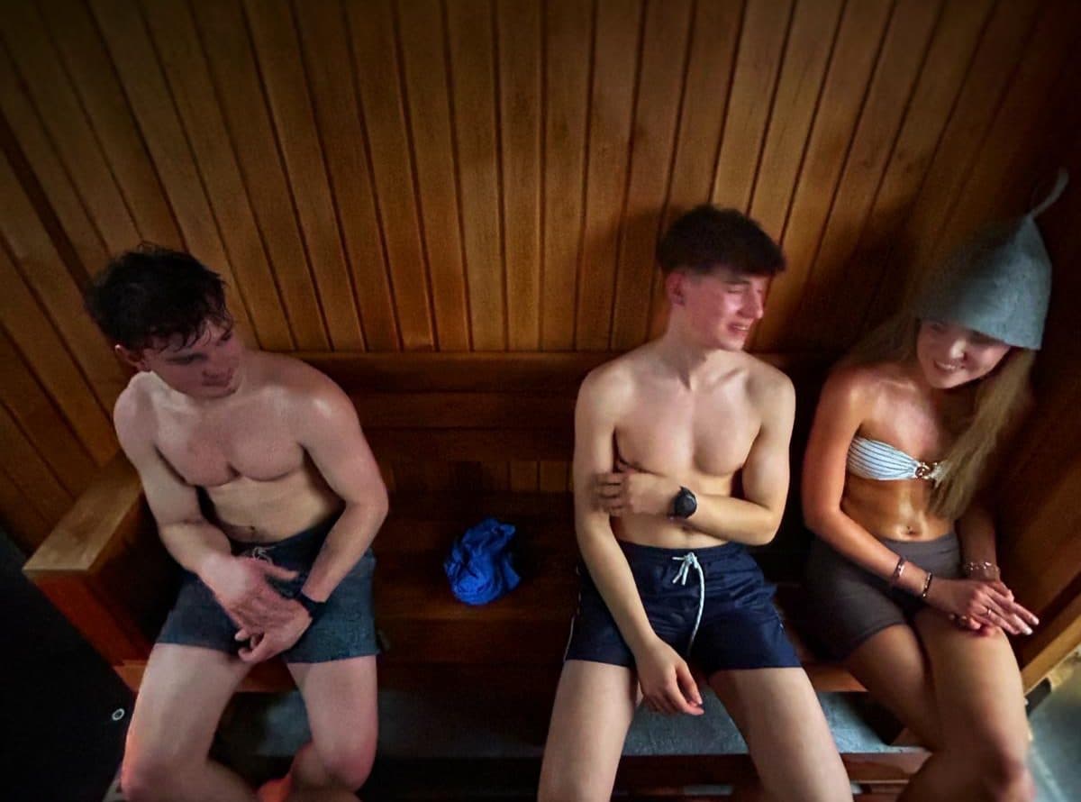 Three young people laughing together on timber benches inside a wood-lined sauna