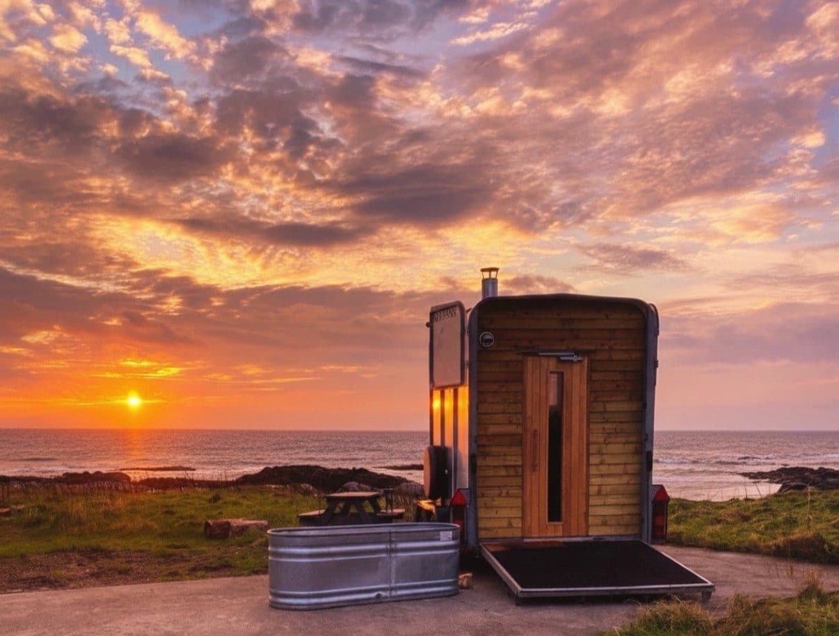 Horsebox sauna with timber cladding and galvanised cold plunge tub on a coastal clifftop at sunset
