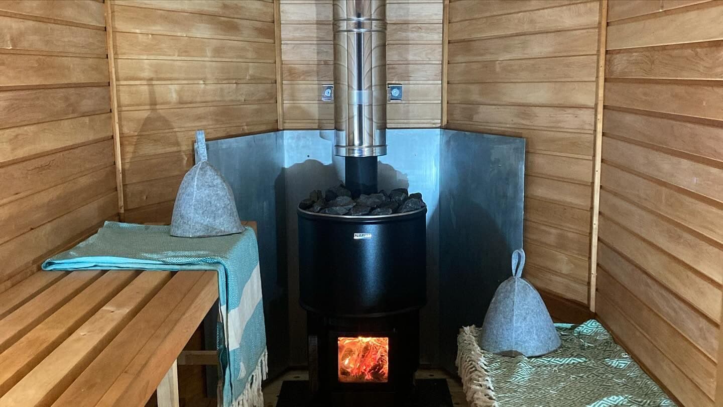 Inside a horsebox sauna showing timber benches, sauna hat, and wood-fired stove with metal heat shield