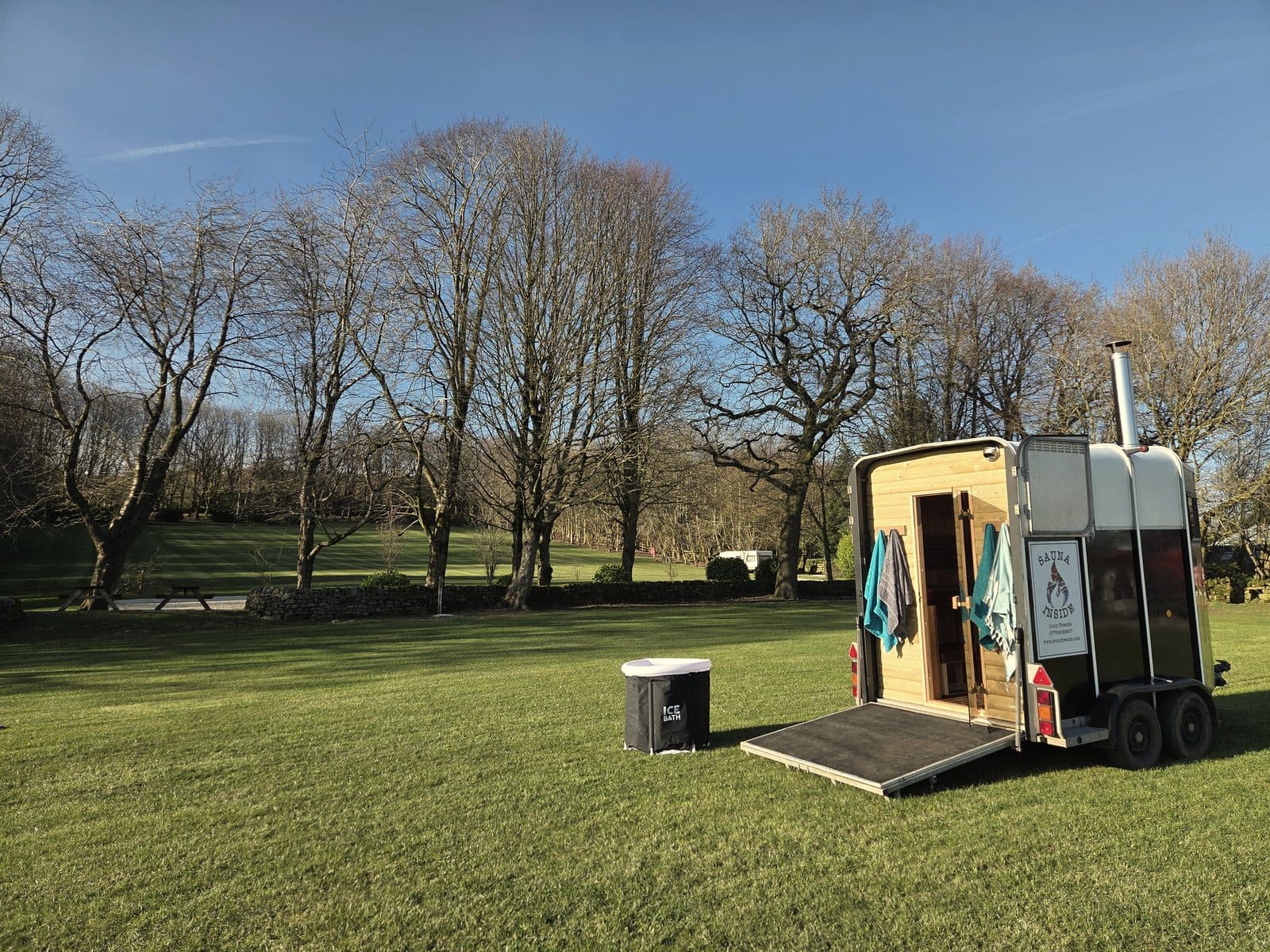 Horsebox sauna with timber interior and ice bath set up on green lawn with bare winter trees and woodland behind