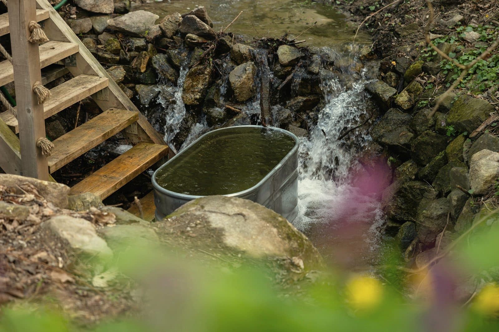 Galvanised stock tank fed by a natural stream among rocks, used as an outdoor cold plunge