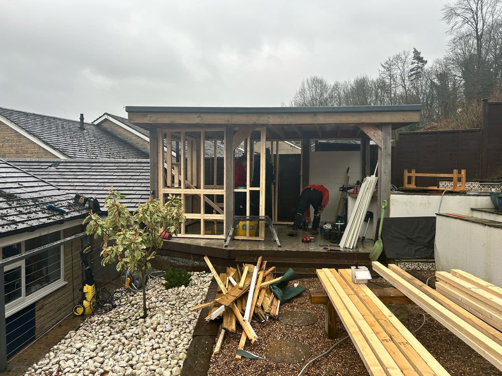 Two builders constructing a timber-framed garden sauna with tools and materials on site